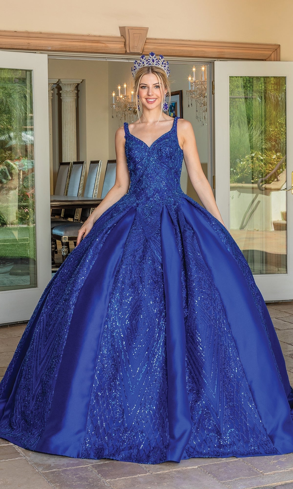 A woman wearing the Quinceanera Dress 1680 By Dancing Queen, featuring a royal blue ball gown with a sweetheart neckline and matching crown, smiles at the camera in front of glass doors.